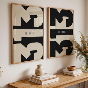 Puede incluir: Dos impresiones artísticas enmarcadas con una combinación de colores negro y crema. Cada impresión presenta la palabra "DETROIT" en un rectángulo horizontal color crema, con grandes formas abstractas crema y negras. Los marcos son de madera clara.