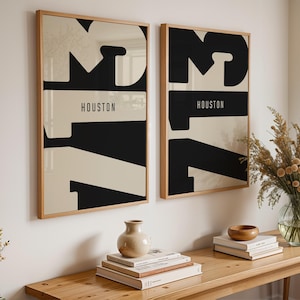 May include: Two framed art prints with a minimalist design. Each print features the word "HOUSTON" in a horizontal black rectangle, with large black and beige abstract shapes. The frames are light wood.