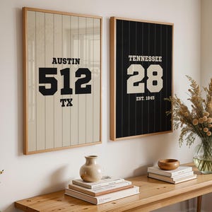 May include: Two framed wall art pieces. One features "Austin 512 TX" in black text on a beige background with vertical lines. The other displays "Tennessee 28 EST. 1845" on a black background with vertical lines. Both are in wooden frames.