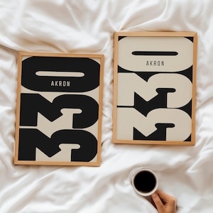 May include: Two framed art prints with the word "AKRON" above the numbers "330" in a bold, modern font. The prints are in a light wood frame, with a black and cream color scheme. A hand holds a cup of coffee.