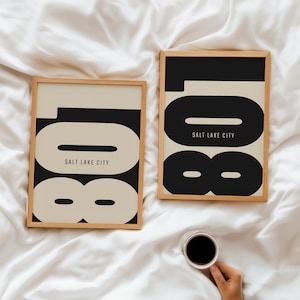 Könnte beinhalten: Zwei gerahmte Kunstdrucke mit minimalistischem Design. Jeder Druck zeigt die Zahl "108" in Schwarz und Beige, darunter "SALT LAKE CITY". Die Drucke werden auf einer weißen Oberfläche präsentiert. Eine Hand hält eine Tasse Kaffee.