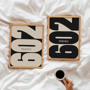 May include: Two framed art prints featuring the number "602" in a bold, black font on a cream background. The word "PHOENIX" is centered below the number. The frames are light wood. A cup of coffee is held in the lower right corner.