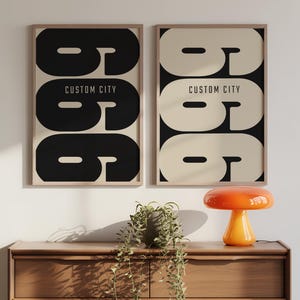 May include: Two framed art prints with a retro design. Each print features a large, stylized number six in black or cream, with the text "CUSTOM CITY" below. The prints are displayed on a wooden chest of drawers.