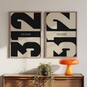 Puede incluir: Dos láminas enmarcadas con un diseño minimalista. Cada lámina presenta el número "23" en negro y crema, con "CHICAGO" en una banda horizontal negra. Las láminas se exhiben sobre una cómoda de madera con una lámpara naranja decorativa.