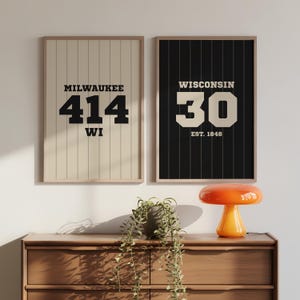 May include: Two framed prints with a vintage baseball jersey design. One features "MILWAUKEE 414 WI" in black on a cream background. The other displays "WISCONSIN 30 EST. 1848" in cream on a black background. Both are in wooden frames.