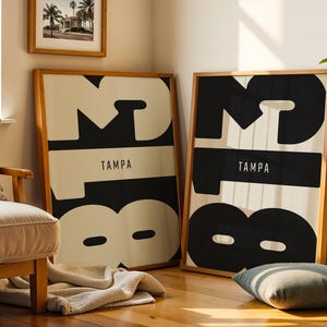 May include: Two framed art prints with a minimalist design. Each print features the word "TAMPA" in a black rectangle, with large black and white abstract numbers. The frames are made of wood.