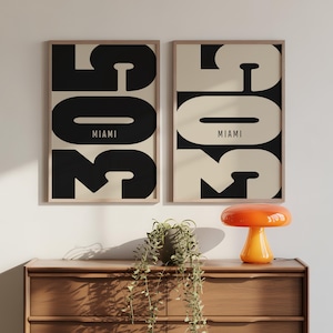 Puede incluir: Dos láminas enmarcadas con el texto "305 MIAMI" en una fuente audaz y moderna. Una lámina tiene texto negro sobre fondo crema, y la otra texto crema sobre fondo negro. Se exhiben sobre una cómoda de madera.