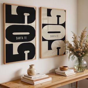 May include: Two framed art prints with a bold black and cream design, featuring the text "SANTA FE". The prints are displayed on a wall above a wooden table with decorative items, including a vase of dried flowers.