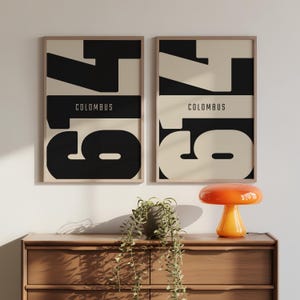 May include: Two framed art prints with a minimalist design. Each print features a large black number 79 against a cream background, with the word "COLUMBUS" in a black horizontal bar. The frames are a light wood colour.