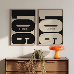 Könnte beinhalten: Zwei gerahmte Kunstdrucke mit minimalistischem Design. Jeder Druck zeigt das Wort "Memphis" in einem Schwarz-Weiß-Farbschema. Die Drucke werden über einer Holzkommode mit einer dekorativen orangefarbenen Lampe angezeigt.