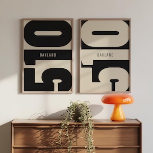 Könnte beinhalten: Zwei gerahmte Kunstdrucke mit der Nummer "510" in großen schwarzen und cremefarbenen Blöcken, mit "OAKLAND" in einem horizontalen Band. Die Drucke werden über einer Holzkommode mit einer orangefarbenen Pilzlampe und einer Topfpflanze ausgestellt.