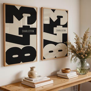 Può includere: Due stampe artistiche incorniciate con un design astratto nero e crema. Le stampe presentano la parola "Charleston" e grandi numeri. Le cornici sono in legno chiaro. Le stampe sono appese a una parete bianca.