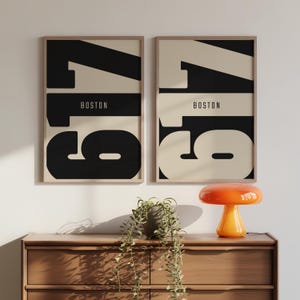 May include: Two framed art prints with the word "BOSTON" and the number "179" in a bold, modern design. The prints feature a black and cream color scheme. A small orange lamp and a plant sit on a wooden dresser below.