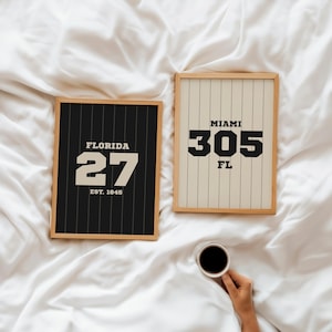 Puede incluir: Dos impresiones enmarcadas con diseños deportivos. Una presenta "FLORIDA 27 EST. 1845" sobre un fondo negro con rayas verticales blancas. La otra muestra "MIAMI 305 FL" sobre un fondo crema con rayas verticales. Una mano sostiene una taza de café.