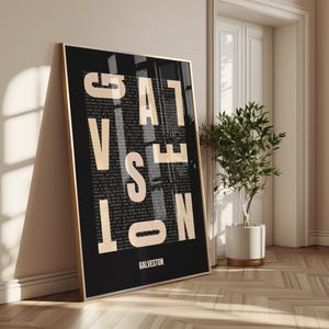 Peut inclure: Une affiche encadrée avec le mot "GALVESTON" en bas. L'œuvre présente les mots "GALVESTON" en grandes lettres dorées en gras, avec un texte plus petit remplissant l'arrière-plan. Le cadre est doré et l'affiche est appuyée contre un mur.