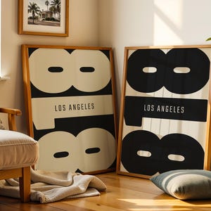 Puede incluir: Dos láminas enmarcadas con un diseño gráfico en blanco y negro. El diseño presenta el número 8 y el texto "LOS ANGELES". Los marcos son de madera marrón claro.