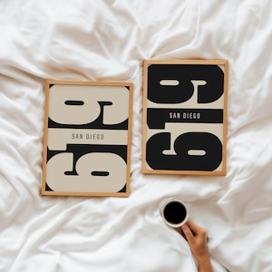 May include: Two framed art prints with the number "619" in a bold, black and cream design, with "SAN DIEGO" printed below. The prints are displayed on a white, textured surface. A hand holds a cup of coffee.