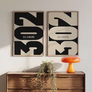 May include: Two framed art prints with the number "302" in black and the word "DELAWARE" in white on a beige background. The prints are displayed above a wooden dresser with a plant and an orange lamp.
