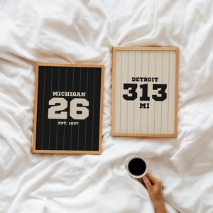Puede incluir: Dos impresiones enmarcadas con diseños deportivos. Una presenta "MICHIGAN 26 EST. 1837" sobre un fondo negro con rayas blancas. La otra dice "DETROIT 313 MI" sobre un fondo crema con líneas verticales. Una taza de café se sostiene en la parte inferior derecha.