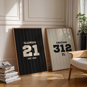May include: Two framed art prints with wooden frames. One print features "ILLINOIS 21 EST. 1818" on a black background. The other print displays "CHICAGO 312 IL" on a cream background. Both prints are leaning against a wall.