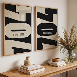 May include: Two framed art prints with a bold black and cream graphic design featuring the number 704 and the name "Charlotte". The prints are displayed on a white wall above a wooden table with decorative items.