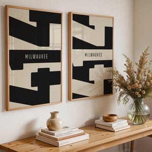 May include: Two framed art prints with a geometric design in black and beige, with the word "MILWAUKEE" in white. The prints are displayed on a white wall above a wooden table with decorative items.