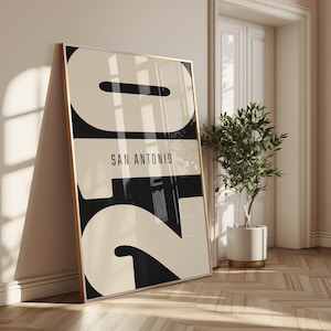 Puede incluir: Un póster enmarcado con un diseño minimalista y audaz que muestra el texto "SAN ANTONIO" en una fuente moderna. La obra de arte utiliza una combinación de colores negro y crema. El póster está apoyado contra una pared en una habitación con suelo de madera y una planta en maceta.