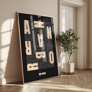 Könnte beinhalten: Ein gerahmter schwarz-goldener Kunstdruck, der an einer Wand lehnt. Der Druck zeigt den Namen "ANN ARBOR" in großen, goldenen Großbuchstaben, wobei kleinerer Text den Hintergrund ausfüllt. Der Text "ANN ARBOR" ist auch am unteren Rand des Rahmens aufgedruckt.
