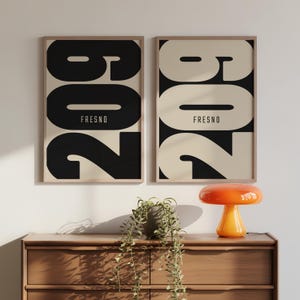 May include: Two framed art prints featuring the number "209" in a bold, black font against a cream background, with "FRESNO" below. The prints are displayed on a wooden dresser, with an orange lamp and a potted plant.