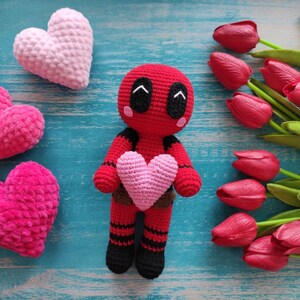 May include: A red crocheted amigurumi toy shaped like a superhero with a pink heart in its hands. The toy is surrounded by pink and red crocheted hearts and a bouquet of red tulips.