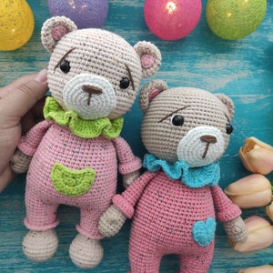 以下が含まれることがあります： 2体の編みぐるみテディベア。1体はピンクで緑と青のアクセント、もう1体はピンクで青のアクセント。両方のクマは茶色の目をしていて、青い表面に座っています。