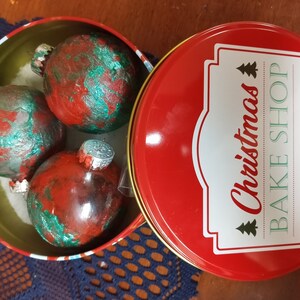 May include: Three red and green Christmas ornaments in a red tin with the words "Christmas Bake Shop" on the lid.