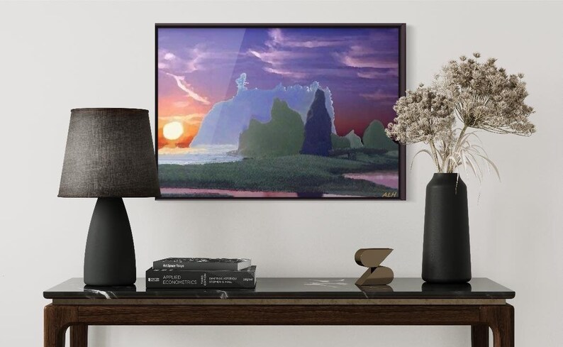 May include: A framed abstract landscape painting with a purple, pink, and blue sky, green and blue rock formations, and a bright orange sun setting over a body of water. The painting is hanging on a white wall above a dark brown wooden table with a black lamp, a vase with dried flowers, and a stack of books.