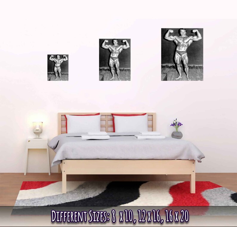 Op de afbeelding: Zwart-witfoto van een bodybuilder die poseert met zijn armen omhoog. De foto is ingelijst en hangt aan een witte muur boven een bed met witte beddengoed. De tekst "DIFFERENT SIZES: 8 X 10, 12 X 16, 16 X 20" is onder de afbeelding gedrukt.