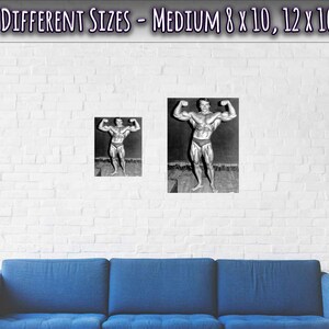 Op de afbeelding: Een zwart-witfoto van een bodybuilder die poseert met zijn armen omhoog. De foto is ingelijst en hangt aan een witte bakstenen muur boven een blauwe bank.