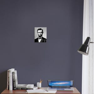 a desk with a laptop, books, and a lamp. A framed black and white portrait of Abraham Lincoln is hanging on the wall above the desk.