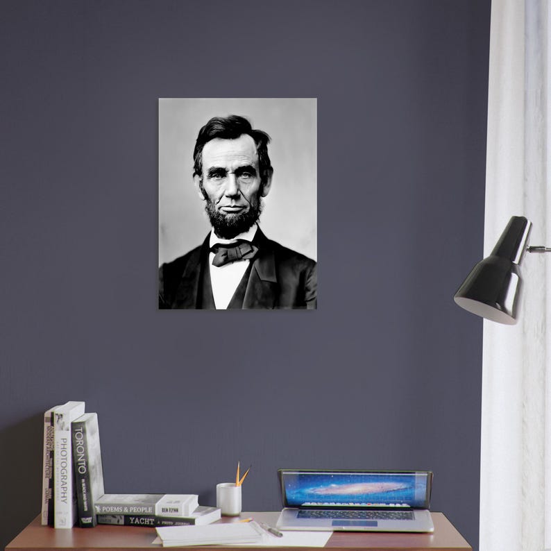 a portrait of Abraham Lincoln hanging on a wall above a wooden desk with a laptop, books, and a lamp.