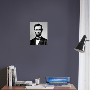 a portrait of Abraham Lincoln hanging on a wall above a wooden desk with a laptop, books, and a lamp.