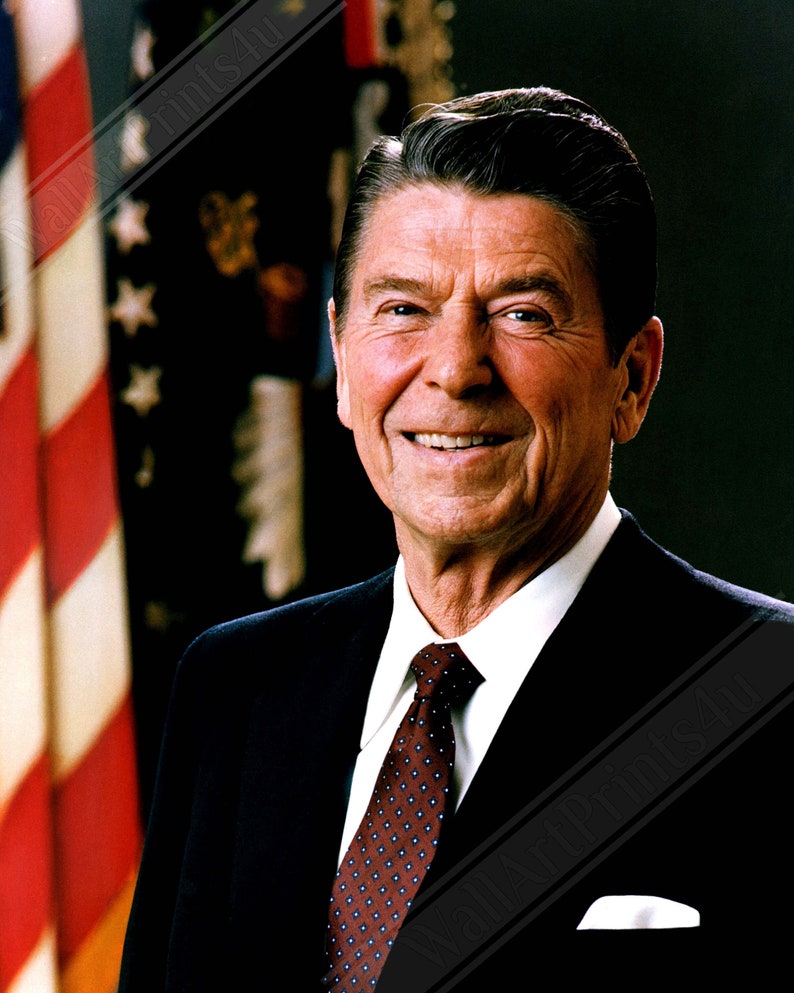 May include: A portrait of a man in a dark suit and a red patterned tie. He is smiling and has a white shirt on. The background is a blurred image of an American flag.