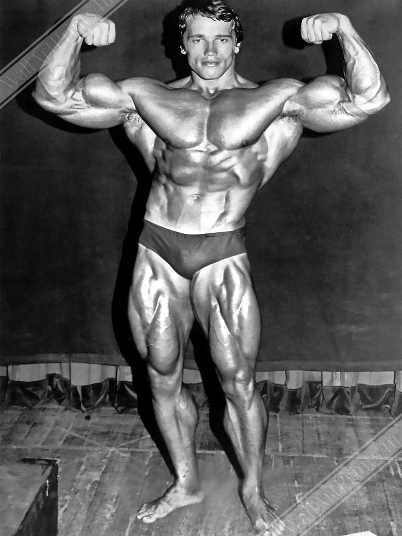 Op de afbeelding: Een zwart-witfoto van een bodybuilder die zijn biceps en quadriceps aanspant. Hij draagt een donkerkleurig zwempak.