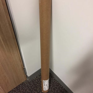 Puede incluir: Un tubo de cart&oacute;n marr&oacute;n con una etiqueta blanca y texto negro. El tubo est&aacute; apoyado contra una pared blanca en una esquina.