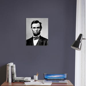 a portrait of Abraham Lincoln hanging on a wall above a wooden desk with a laptop, books, and a lamp.