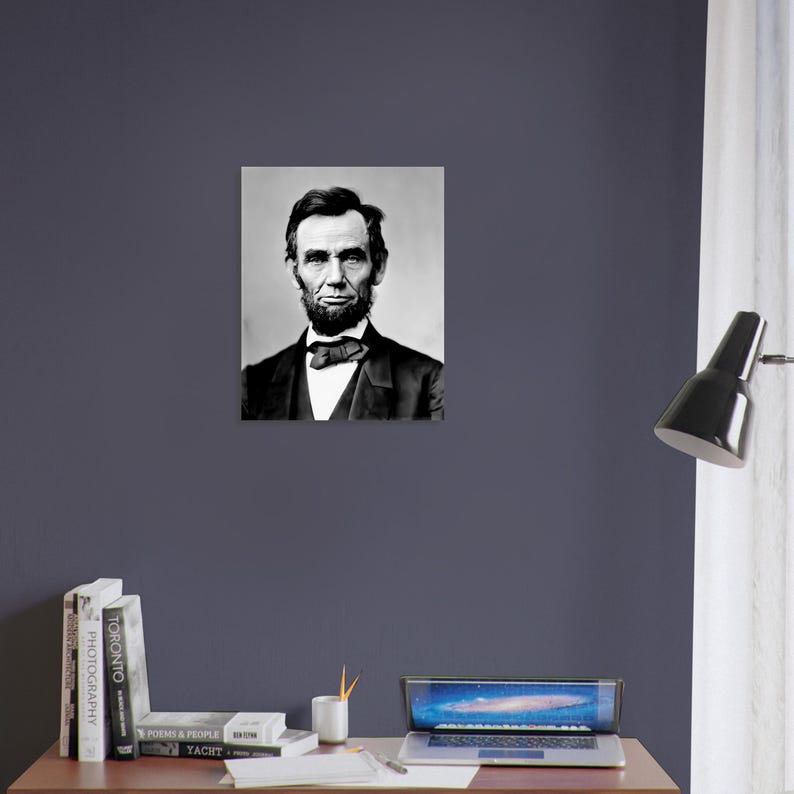 a portrait of Abraham Lincoln hanging on a wall above a wooden desk with a laptop, books, and a lamp.