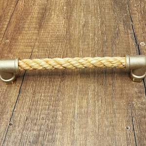 Può includere: Una maniglia orizzontale per cassetto caratterizzata da una sezione centrale in corda intrecciata color beige. La corda è fissata alle due estremità da raccordi per tubi in metallo color oro opaco a forma di gomito, creando un elemento robusto.