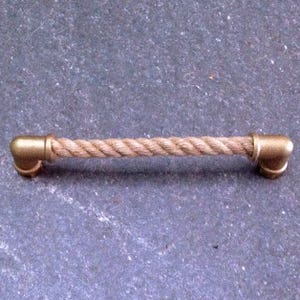 Op de afbeelding: Een horizontale ladehandgreep met een middengedeelte van dik, gedraaid beige touw. Het touw is aan beide uiteinden bevestigd met metalen, goudkleurige L-vormige buiskoppelingen die dienen als stevige bevestigingspunten voor de handgreep.