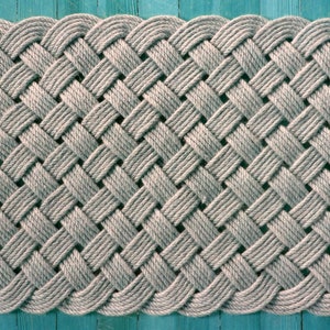 May include: A gray woven doormat with a diamond pattern. The mat is made of thick rope and has a slightly rounded edge.