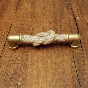 Puede incluir: Un tirador de cajón que presenta una sección central de cuerda trenzada de color beige claro atada en un nudo, conectada en ambos extremos a accesorios de tubería metálicos de color latón en forma de L con acabado mate y bases circulares.