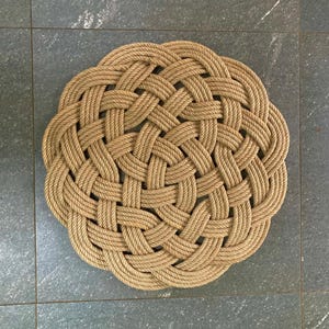 May include: A circular mat made from woven rope, presented in a natural beige hue. The mat features a complex basket-weave design, constructed from thick rope. It measures approximately 76 cm in diameter, suitable for various settings.
