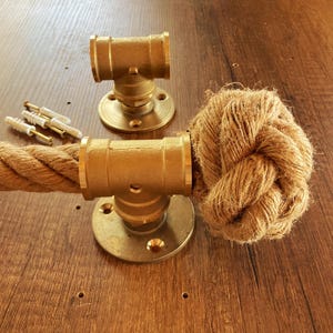 Könnte beinhalten: Ein nautisch inspiriertes Seil- und Messing-Hardware-Set. Das Set enthält ein dickes, naturfarbenes Seil, Messingbeschläge und Montagematerial. Das Seil ist an einem Ende geknotet. Die Messingbeschläge haben eine runde Basis.