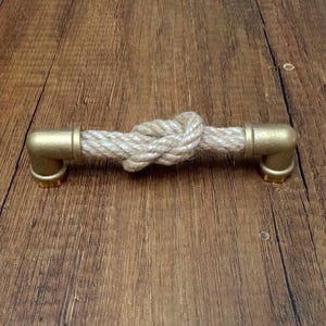 Op de afbeelding: Een ladehandgreep bestaande uit een dik, beige gedraaid touw met een centrale knoop, aan beide uiteinden bevestigd met geborsteld goudkleurige metalen buiskoppelingen. Het beslag is ontworpen voor horizontale montage op een meubelstuk.